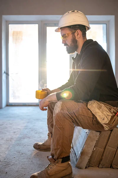A man builder in a helmet and goggles drinks a hot drink. — Stock Photo, Image