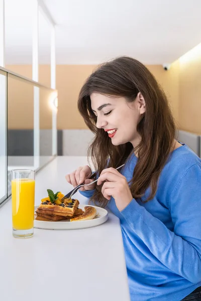 Atractiva joven come gofres belgas y jugo para el desayuno. — Foto de Stock