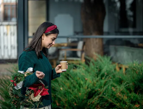 Mujer joven con una corona de Navidad y una taza de café en un paseo por la ciudad. — Foto de Stock