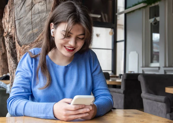 Una joven con auriculares inalámbricos usa el teléfono mientras está sentada en un café. — Foto de Stock