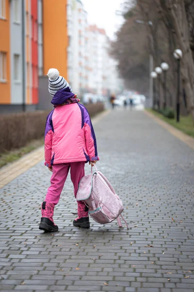 Holčička s batohem, v bundě a klobouku poblíž školy. — Stock fotografie