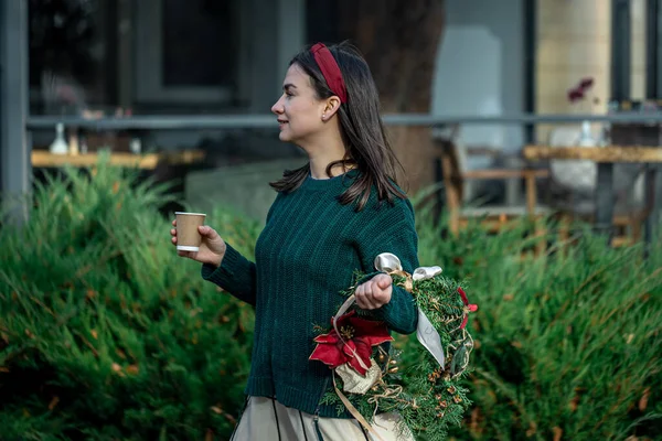 Mladá žena s vánočním věncem a šálkem kávy na procházce po městě. — Stock fotografie