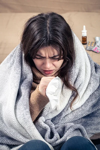En ung kvinde hoster mens hun sidder hjemme, pakket ind i et tæppe. - Stock-foto