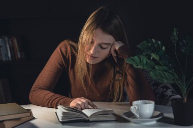 Genç bir kadın masada oturur ve gece lambasıyla kitap okur..