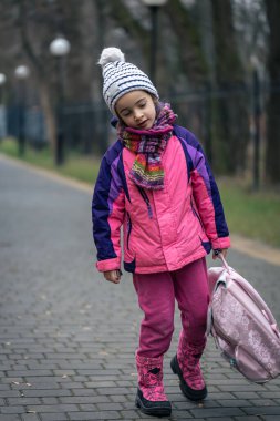 Okul yakınlarında ceketli ve şapkalı sırt çantalı küçük bir kız..
