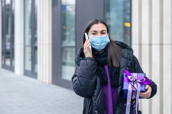 Femeia tânără într-o mască de protecție cu un cadou în mâini vorbind la telefon. — Fotografie, imagine de stoc