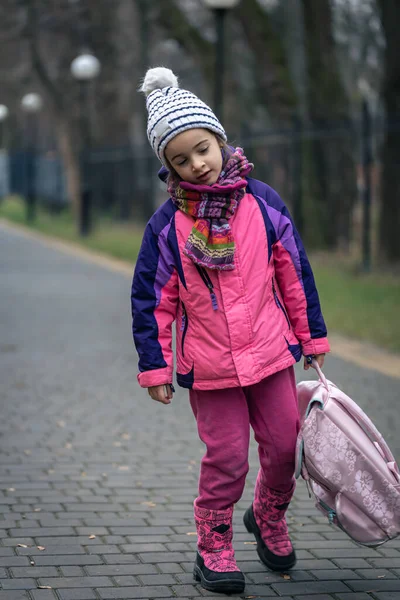 Kleines Mädchen mit Rucksack, Jacke und Hut in der Nähe der Schule. — Stockfoto