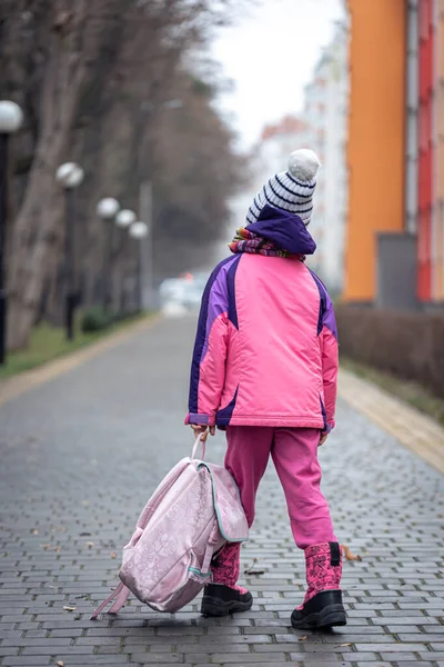 Mała dziewczynka z plecakiem, w kurtce i kapeluszu w pobliżu szkoły.. — Zdjęcie stockowe