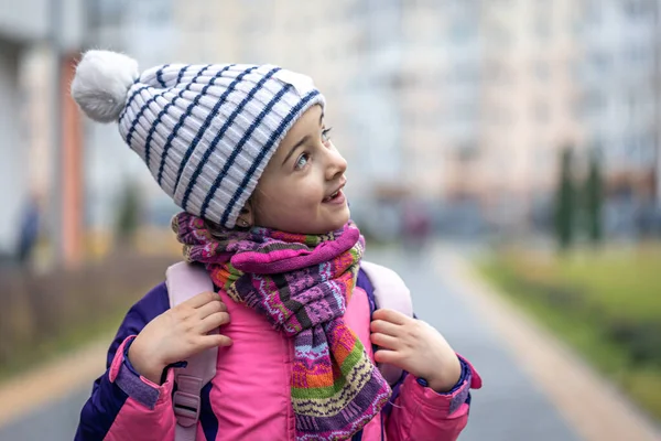Petite fille avec un sac à dos, dans une veste et un chapeau près de l'école, espace de copie. — Photo