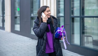 Elinde mor bir hediyeyle dışarıda telefonla konuşan genç bir kadın..