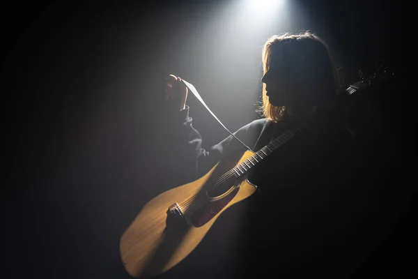 Uma jovem mulher com uma guitarra acústica no escuro sob um raio de luz. — Fotografia de Stock