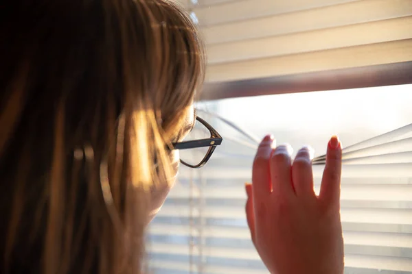 Uma mulher olha através dos blinds na luz solar da manhã adiantada. — Fotografia de Stock