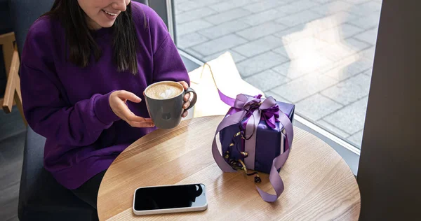 Mujer joven en un suéter púrpura en un café con una taza de café. — Foto de Stock