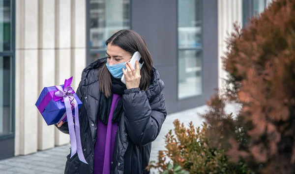 Femeia tânără într-o mască de protecție cu un cadou în mâini vorbind la telefon. — Fotografie, imagine de stoc