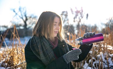 Genç bir kadın, kışın yürüyüş yaparken termostan sıcak bir içecek alır..