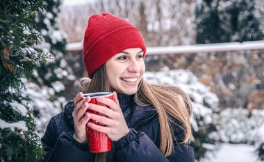 Genç bir kadın kışın kırmızı bir termal bardaktan sıcak bir içecek içer..