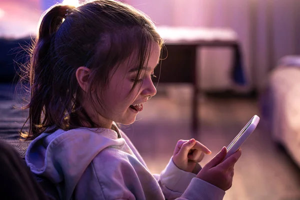 Ein kleines Mädchen benutzt ein Smartphone, während es in ihrem Zimmer sitzt. — Stockfoto