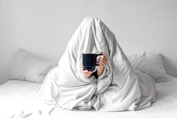 Una mujer envuelta en una manta se sienta en la cama con una taza de café en las manos. — Foto de Stock