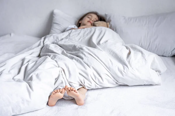 Las niñas pequeñas piernas sobresalen de debajo de la manta blanca. — Foto de Stock