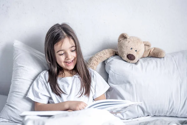 La niña lee un libro con un osito de peluche en la cama por la mañana. —  Fotos de Stock