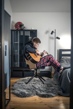 Genç bir adam evindeki odasında akustik gitar çalar..