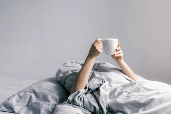 Kobieca dłoń trzyma filiżankę kawy leżąc w łóżku. — Zdjęcie stockowe