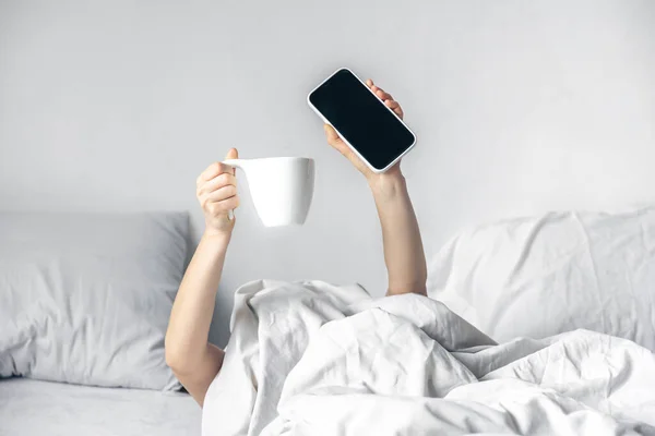 Una mujer se acuesta en la cama con un smartphone y una taza de café en las manos. — Foto de Stock