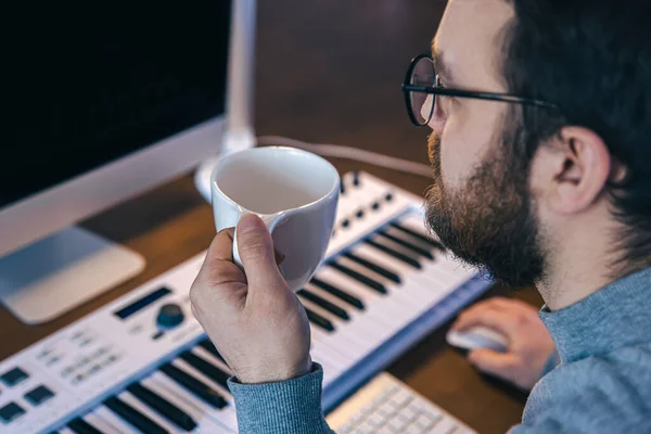 Een man met een kop koffie zit voor een computer en een muzikaal toetsenbord. — Stockfoto