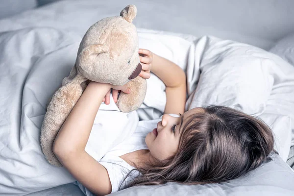 Little cute girl in bed with a teddy bear early in the morning. — Stock Photo, Image