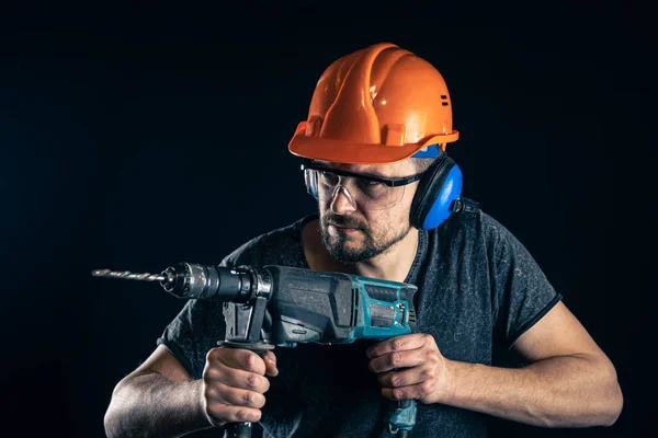 Un joven en mono casco protector casco duro mantenga taladro eléctrico. — Foto de Stock