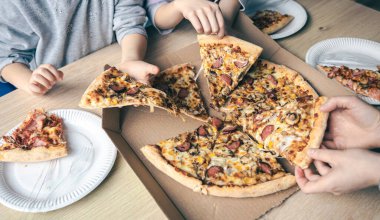 Masadaki karton kutudan sıcak pizza dilimleri alan gençler..