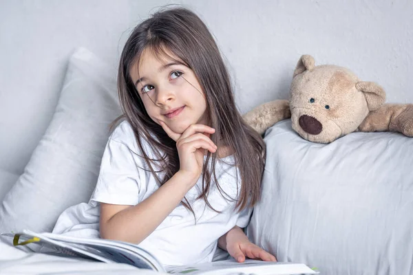 La niña lee un libro con un osito de peluche en la cama por la mañana. —  Fotos de Stock