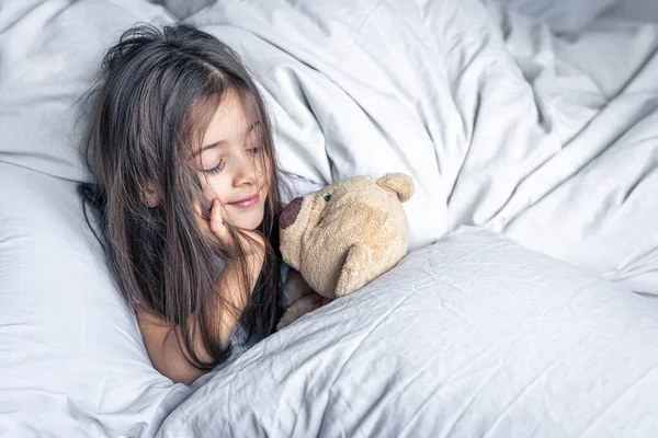 Little cute girl in bed with a teddy bear early in the morning, copy space. — Stock Photo, Image