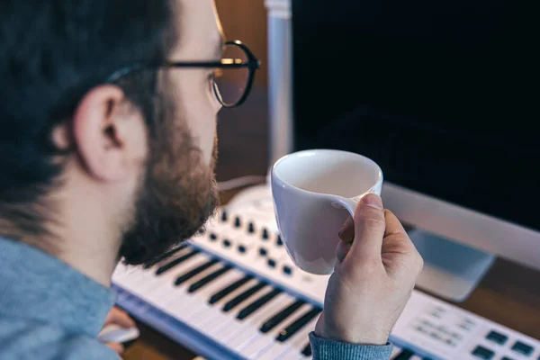 Een man met een kop koffie zit voor een computer en een muzikaal toetsenbord. — Stockfoto
