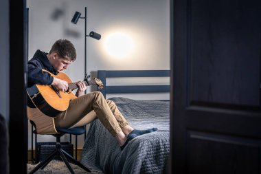 Genç bir adam evindeki odasında akustik gitar çalar..