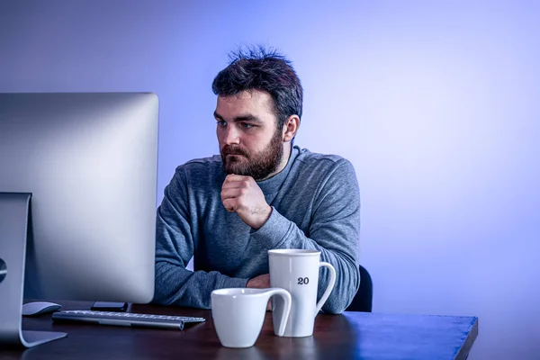 Hombre cansado se sienta frente a una computadora con una taza de café, iluminación de colores. — Foto de Stock
