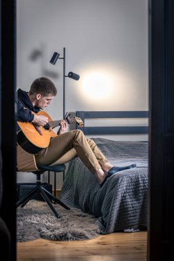 Genç bir adam evindeki odasında akustik gitar çalar..