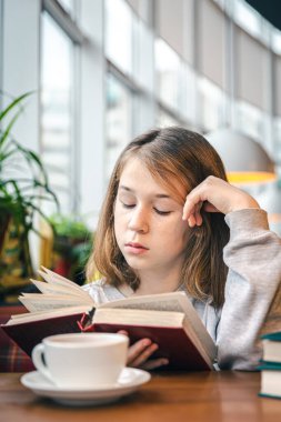 Küçük bir kız bir kafede bir fincan çayla otururken kitap okuyor..