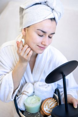 Genç bir kadın yüzüne krem sürüyor, aynaya bakıyor..