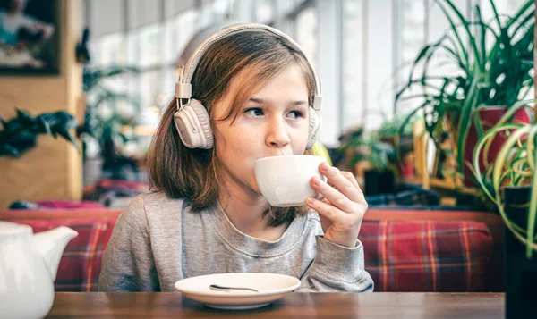 A little girl in headphones in a cafe with a cup of tea. — Stock Photo, Image