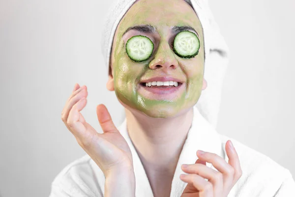 Jonge vrouw met een groen gezichtsmasker en verse komkommers in een witte badjas. — Stockfoto