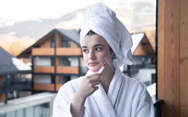 Eine junge Frau mit einem Handtuch auf dem Kopf macht eine Massage mit einer Gesichtsrolle. — Stockfoto