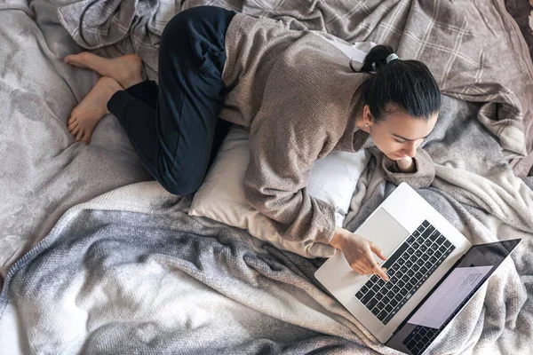 Uma mulher trabalhando em um laptop enquanto deitada na cama, vista superior. — Fotografia de Stock