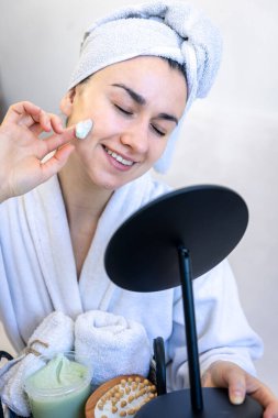 Genç bir kadın yüzüne krem sürüyor, aynaya bakıyor..