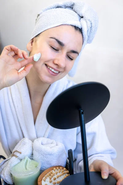 Genç bir kadın yüzüne krem sürüyor, aynaya bakıyor..