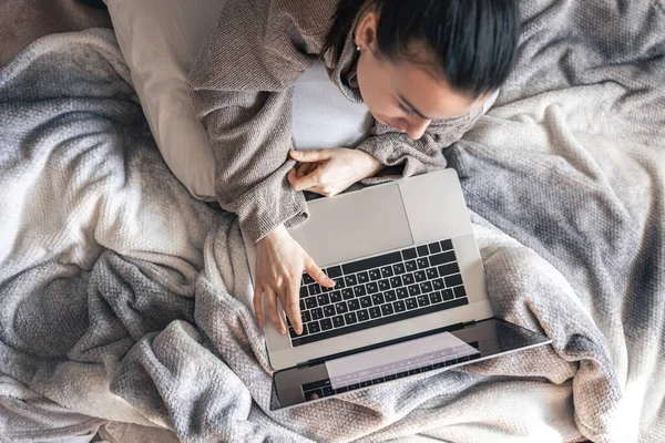 Uma mulher trabalhando em um laptop enquanto deitada na cama, vista superior. — Fotografia de Stock
