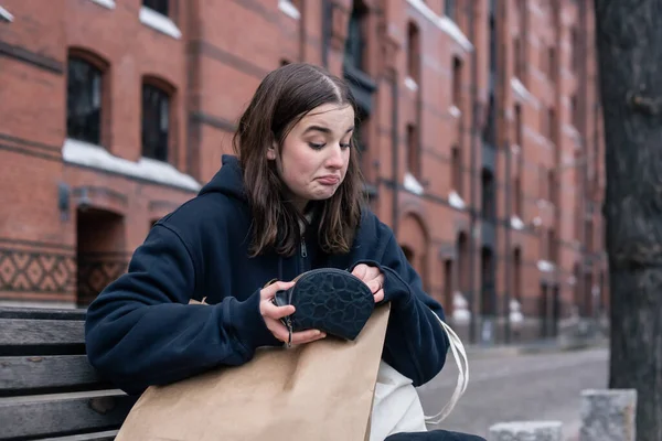 Seorang wanita muda duduk di bangku dengan dompet kosong, konsep kekurangan uang. — Stok Foto