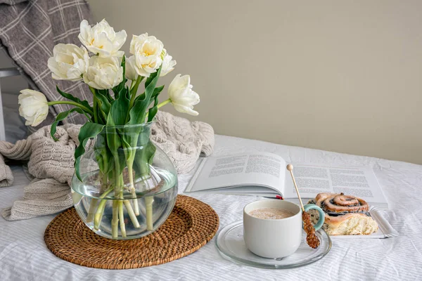 Cozy composition with a bouquet of white tulips, coffee and a bun in bed.