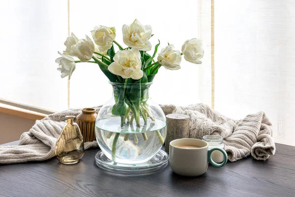Home composition with a bouquet of tulips in a glass vase, a knitted element, a cup of coffee and candles.