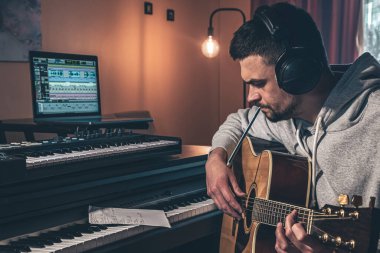 Erkek müzisyen, evde bilgisayar ve piyanonun yanındaki iş yerinde akustik gitar çalar..